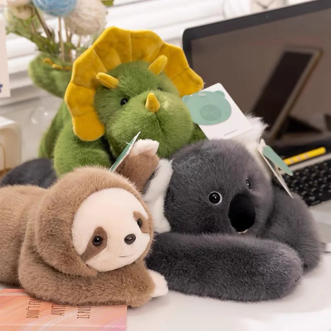 Plush stuffed toys of a green dinosaur, gray koala, and brown sloth on a desk beside a laptop.